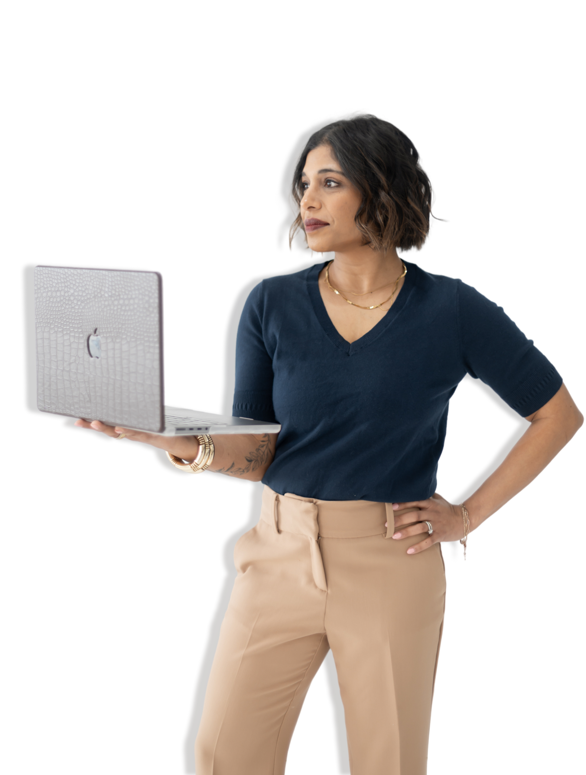 A woman with short dark hair, wearing a navy blue top and beige pants, stands holding an open laptop in one hand and looks to the side against a white background.
