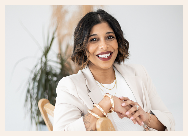 A woman with short dark hair and a bright smile sits on a wooden chair, wearing a light blazer, gold jewelry, and a white top, with a leafy plant in the background.