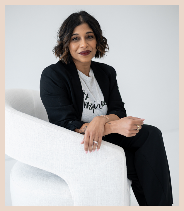 A woman with shoulder-length dark hair and wearing a black blazer and white t-shirt sits on a white modern chair, smiling slightly and looking at the camera in a bright, minimalist setting.