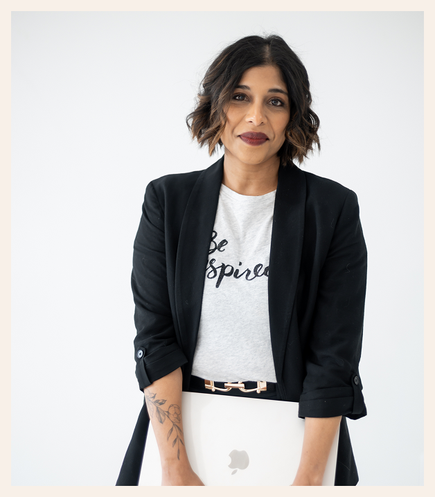 A woman with short wavy hair and a tattoo on her forearm stands against a white background, wearing a black blazer over a T-shirt that says Be Inspired, holding a closed laptop in front of her.