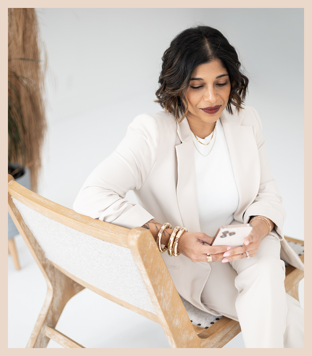 A woman with shoulder-length dark hair sits on a wooden chair, wearing a light cream suit and several gold bracelets, looking down and smiling slightly while holding a smartphone.