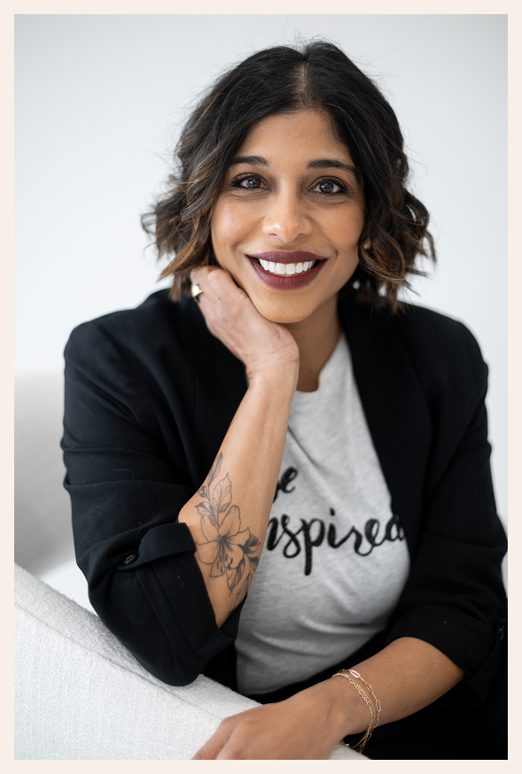 A woman with shoulder-length wavy hair smiles while sitting on a chair. She wears a black blazer, a white shirt that reads be inspired, has a floral tattoo on her forearm, and rests her chin on her hand.