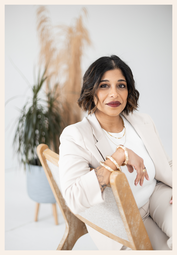 A woman with shoulder-length dark hair and a beige suit sits on a chair, smiling slightly. She wears gold jewelry and poses confidently in a bright room with tall plants in the background.