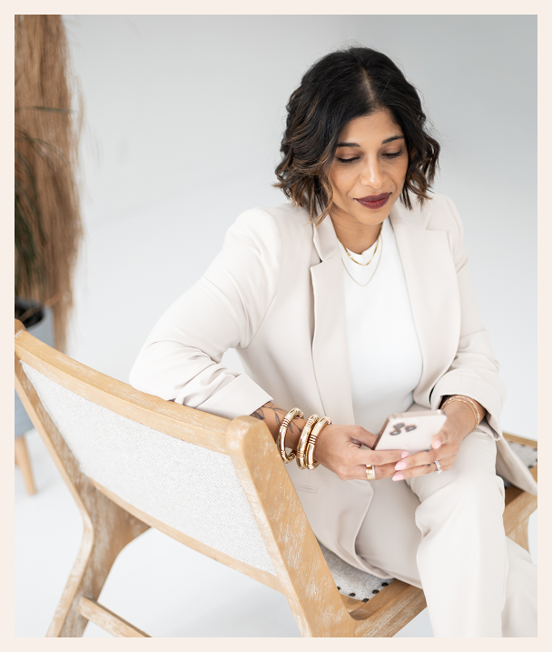 A woman with shoulder-length dark hair, wearing a light-colored suit and multiple bracelets, sits on a wooden chair and looks at her phone with a slight smile in a bright, minimalistic room.