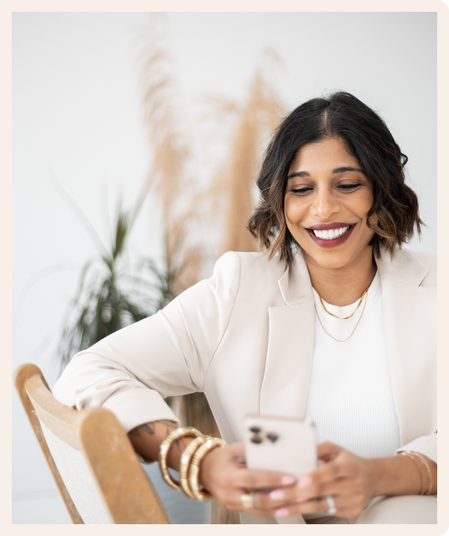 A woman in a beige suit smiles while looking at her phone. She is sitting on a chair in a bright room with plants in the background, exuding a cheerful and relaxed vibe.