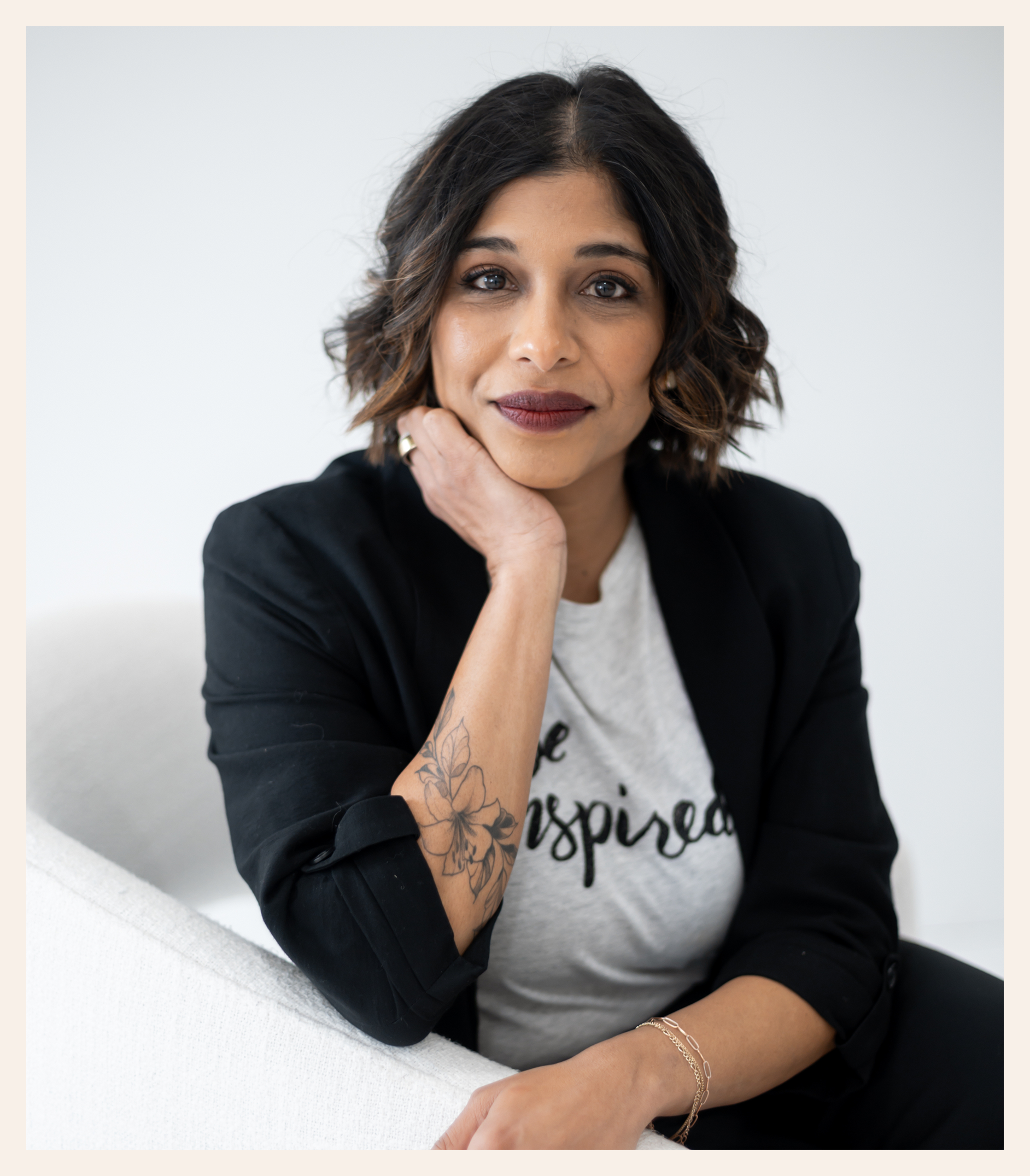 A woman with shoulder-length dark hair and a floral tattoo on her forearm sits on a white chair, wearing a black blazer and a white shirt that reads be inspired, looking confidently at the camera.