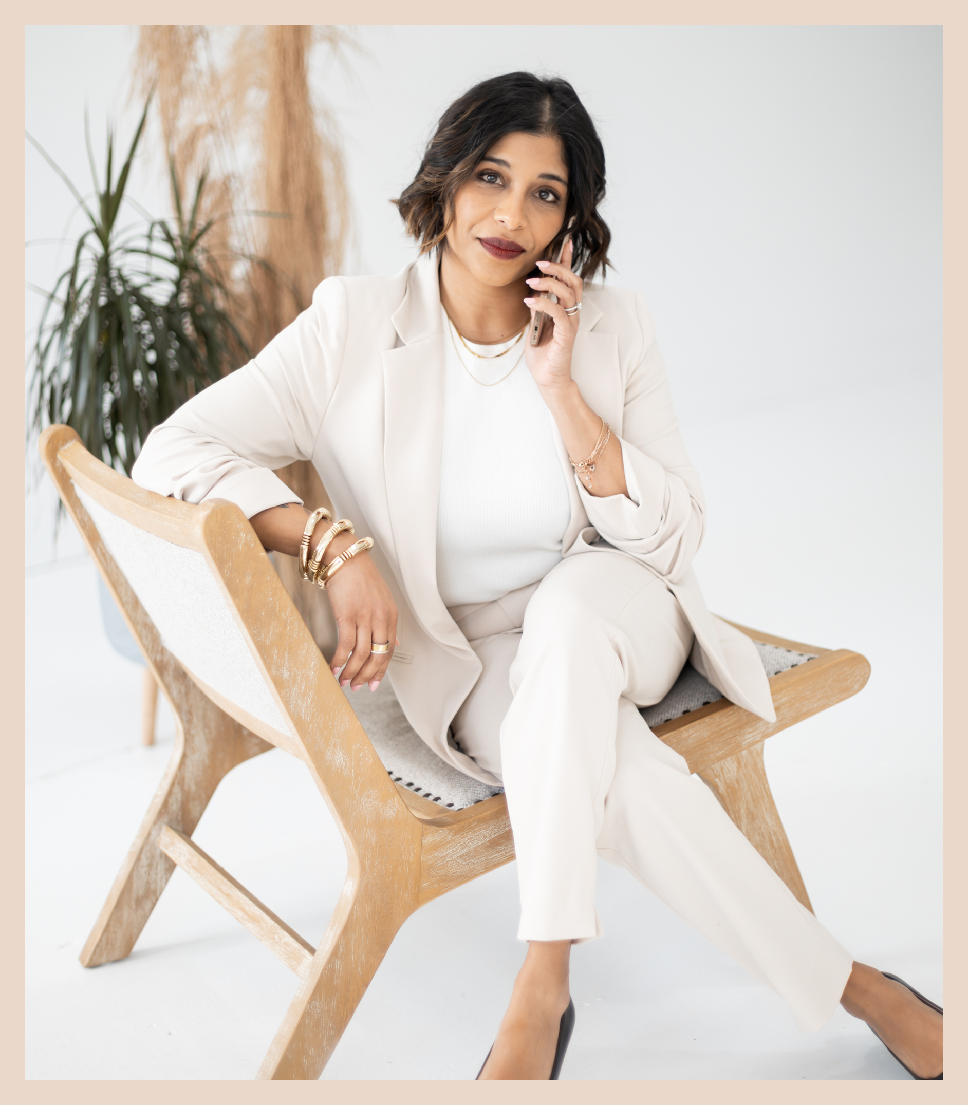 A woman in a cream-colored suit sits on a wooden chair, smiling slightly while talking on her phone. She wears gold jewelry and has shoulder-length dark hair. There are dried plants in the background.