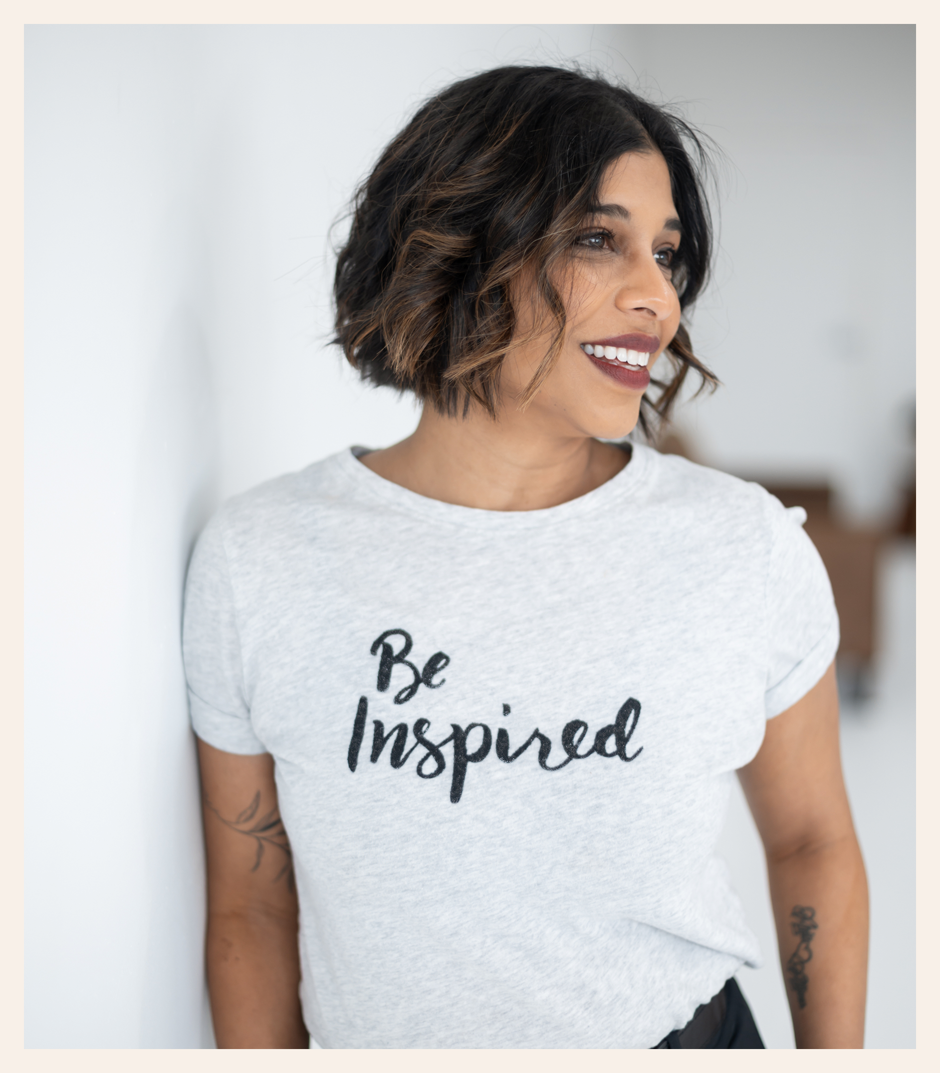 A woman with short wavy hair smiles while leaning against a white wall. She is wearing a light gray t-shirt with the words Be Inspired written on it in black script.